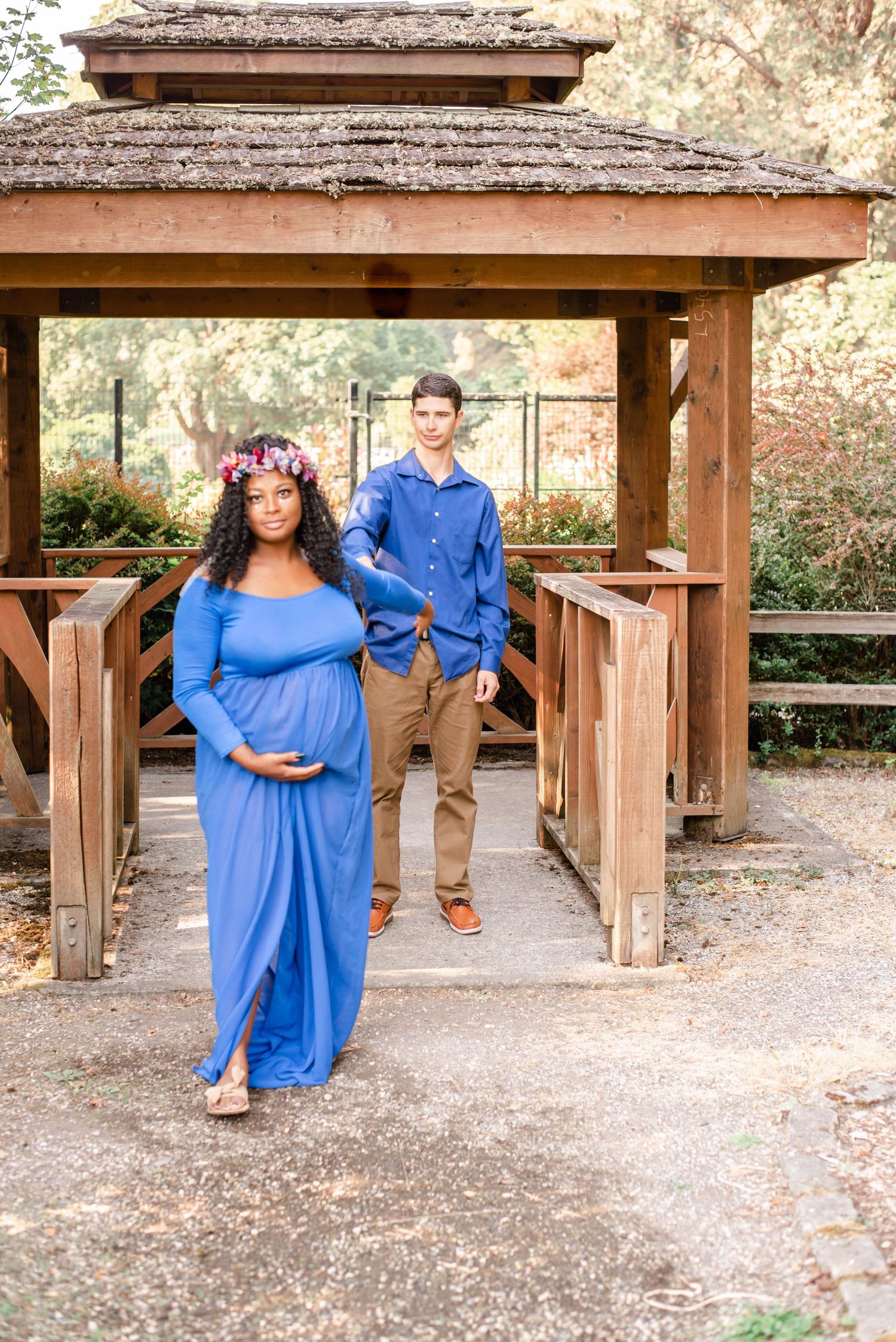 Couple poses under wooden gazebo during outdoor maternity photoshoot.