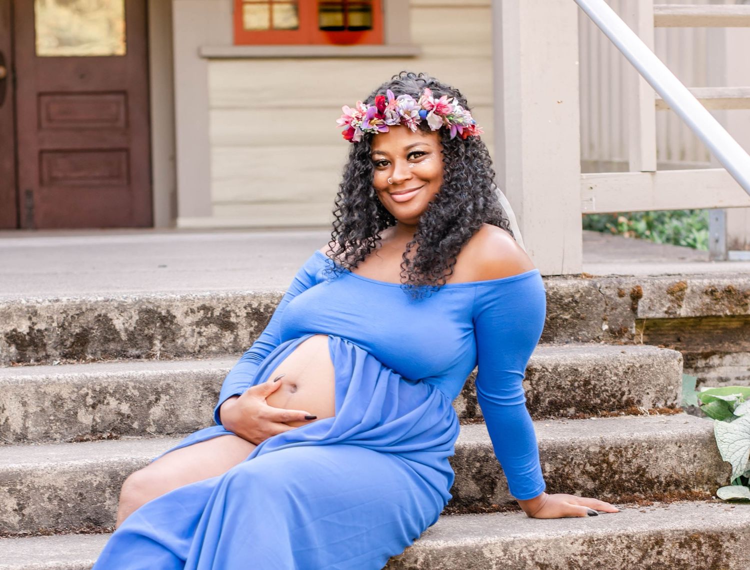 Expecting mother in blue dress and floral crown sits on concrete steps outdoors.