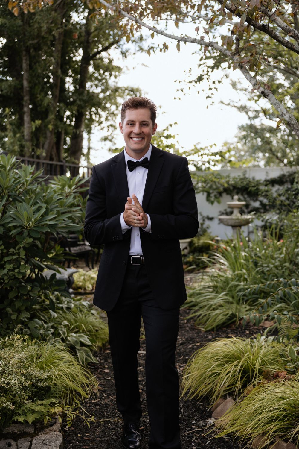 Groom in classic black tuxedo smiles while standing in lush garden pathway.
