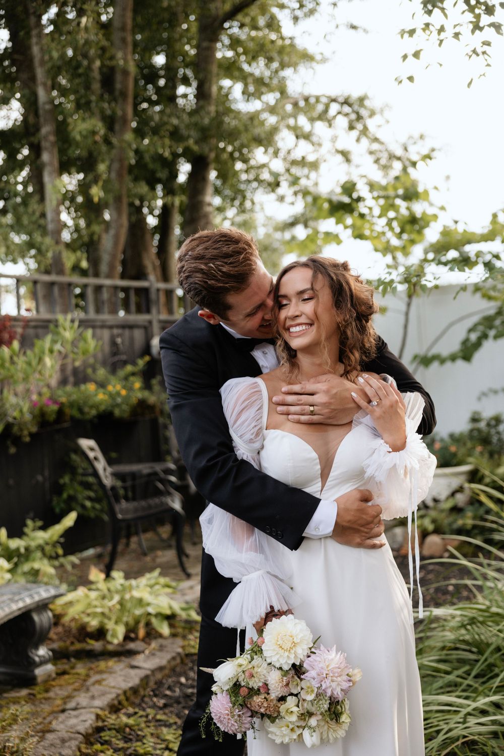 Joyful newlyweds embrace outdoors with bride holding a pastel bouquet in garden setting.