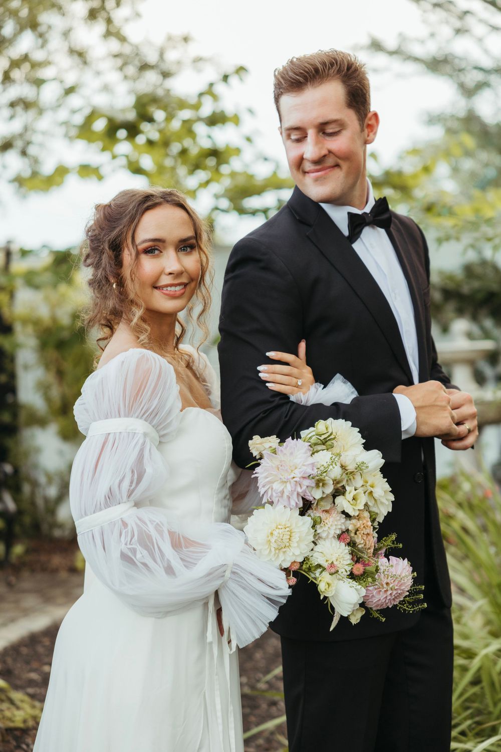 Elegant wedding couple poses together in a garden setting with a romantic bouquet of pink and white flowers.
