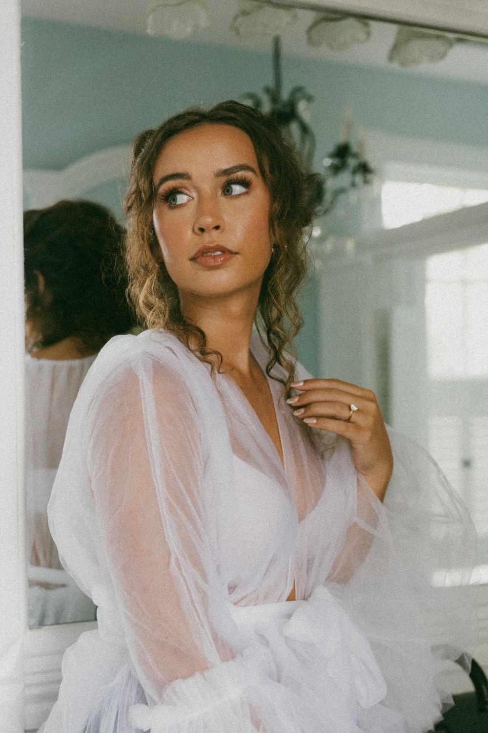 Bride in ethereal sheer white gown poses thoughtfully in bright interior space.