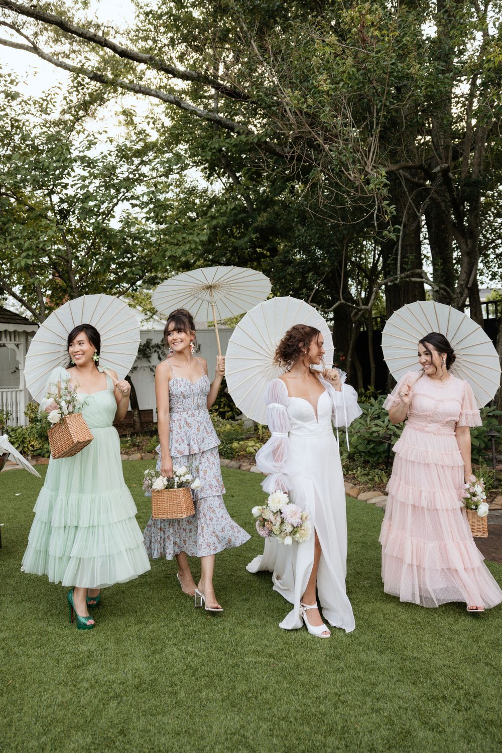 Bridal party holding white parasols poses in pastel dresses on manicured lawn.