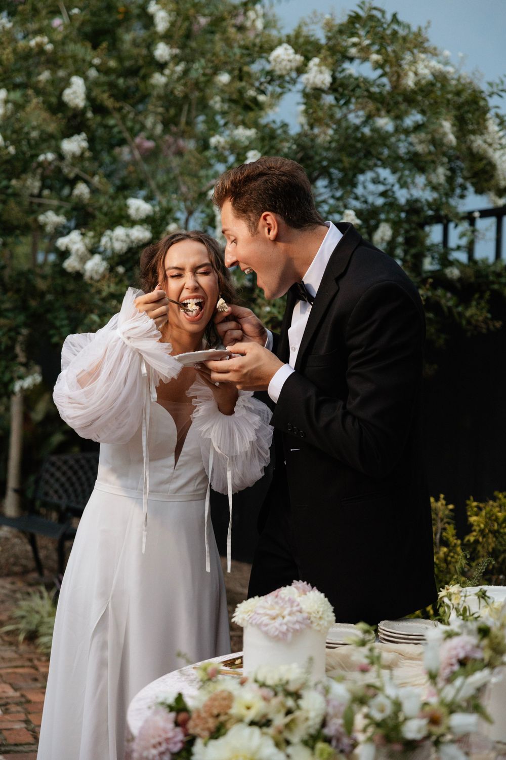 Newlywed couple shares cake and laughter at outdoor reception with floral decorations.