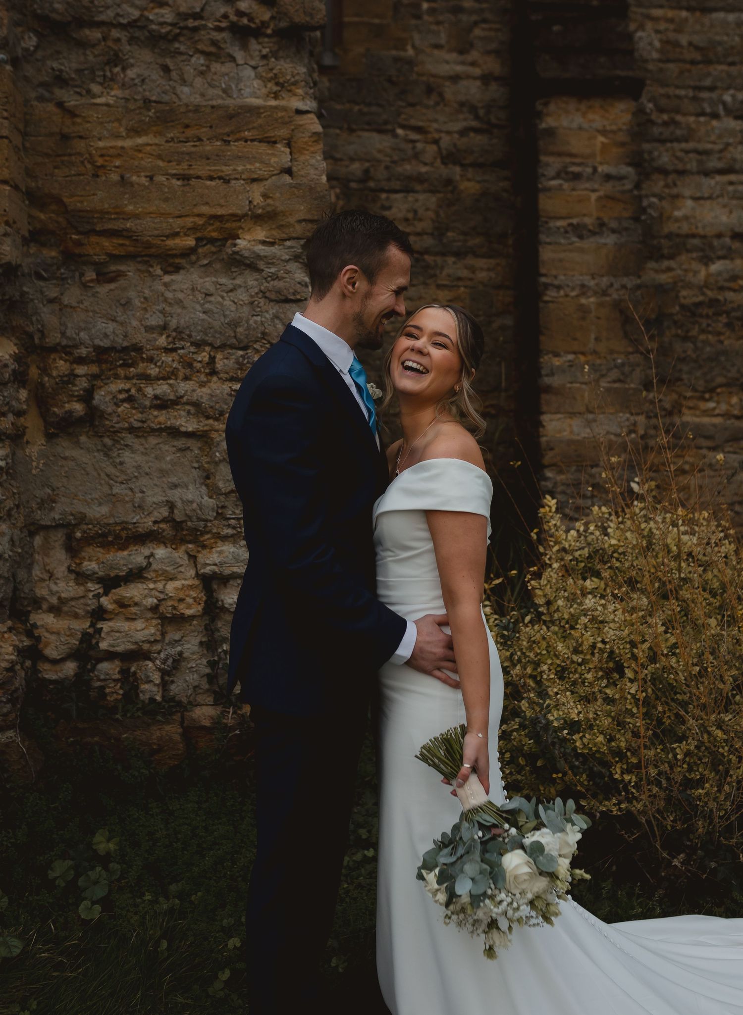 Wedding couple sharing intimate moment against rustic stone wall with bridal bouquet.