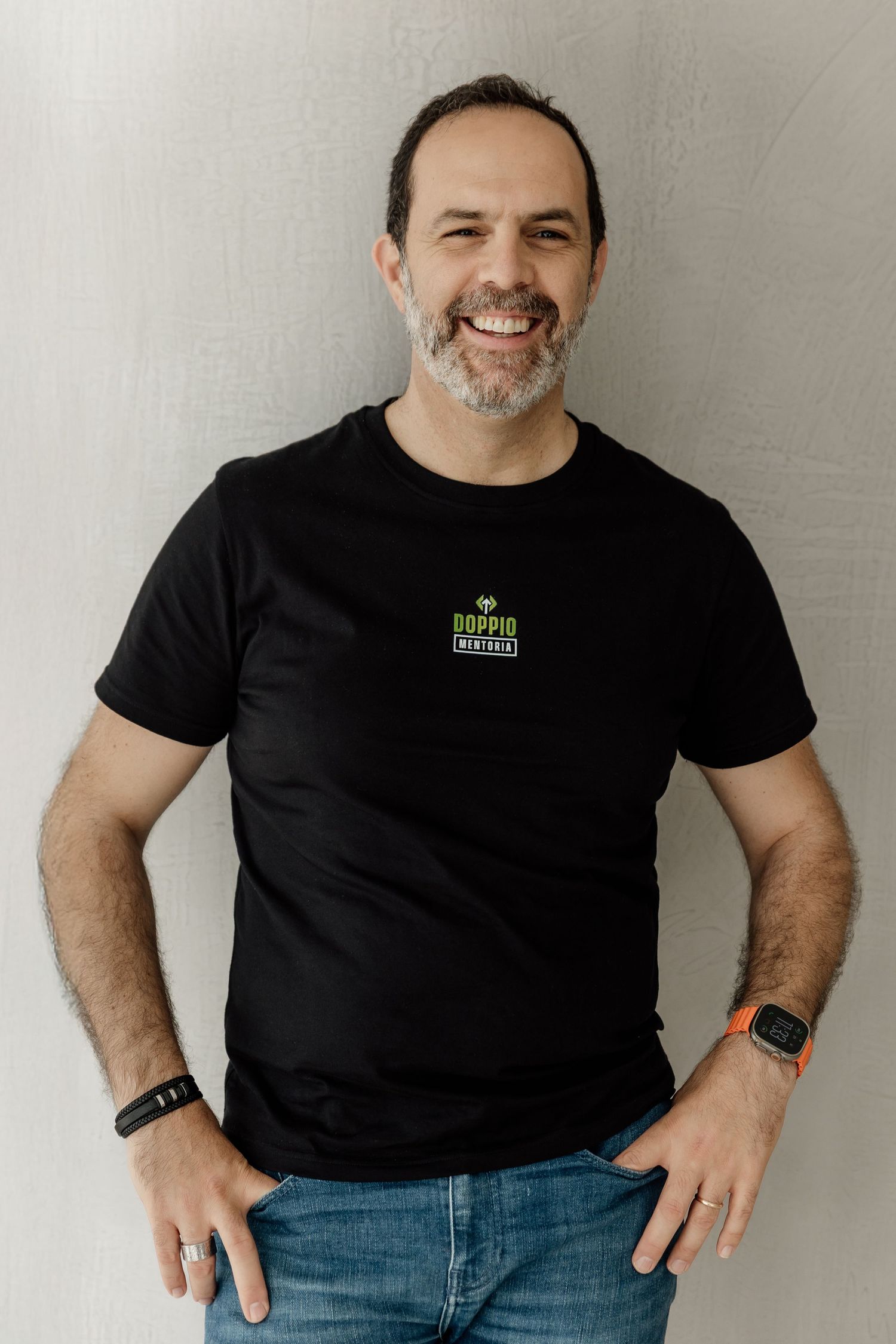 A person in a black t-shirt with a small logo stands against a white wall wearing jeans and a smart watch.