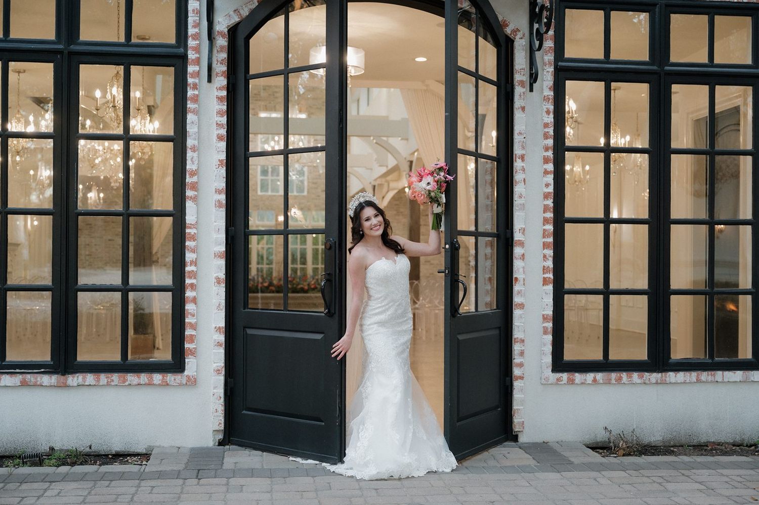 A bride in a white lace gown stands in an elegant doorway with black framed windows and vintage architectural details.