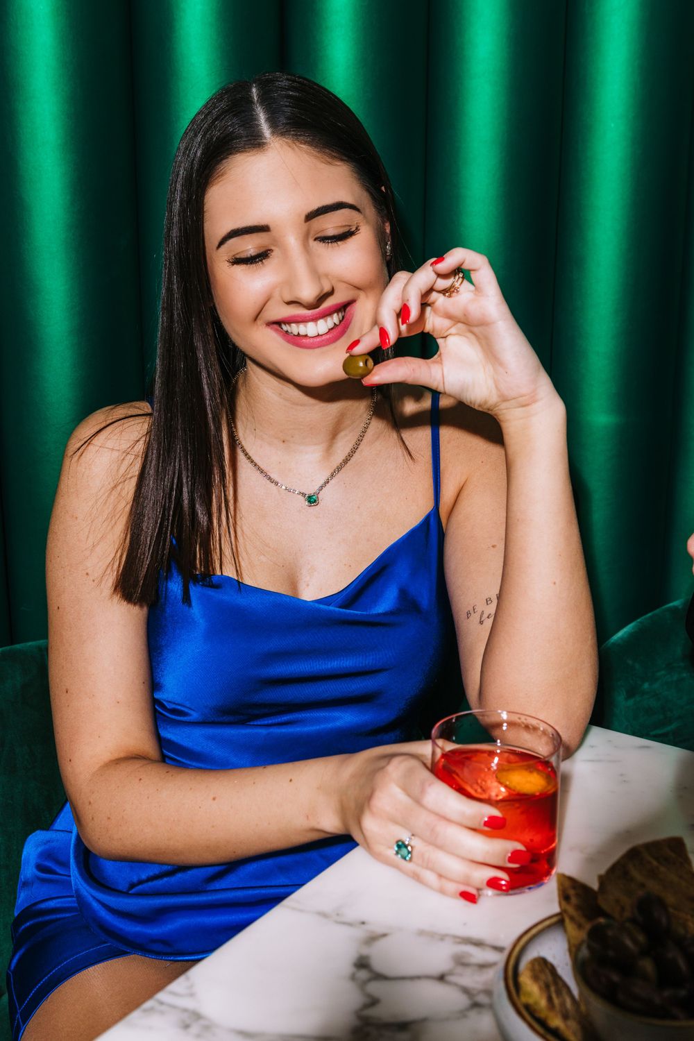 Person in blue satin dress enjoying cocktail against emerald green curtain backdrop.
