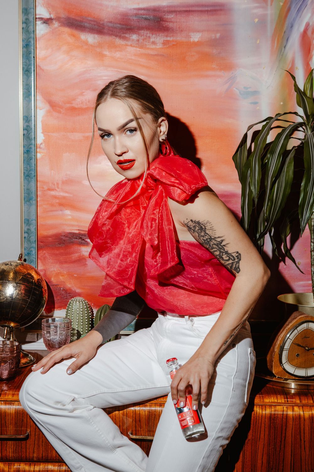 Stylish woman in red top and white pants posing with drink against coral background.