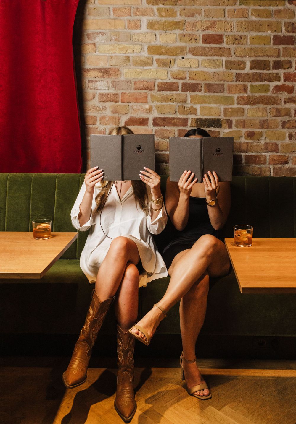 Two people sitting on couch holding books up to cover faces against brick wall backdrop.