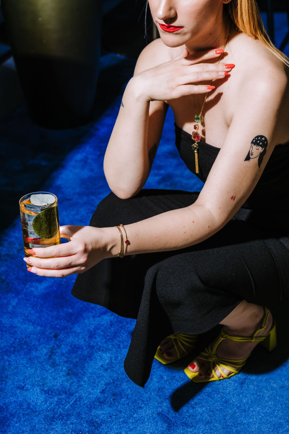 Elegant figure on blue carpet holding cocktail with painted nails and visible tattoo.