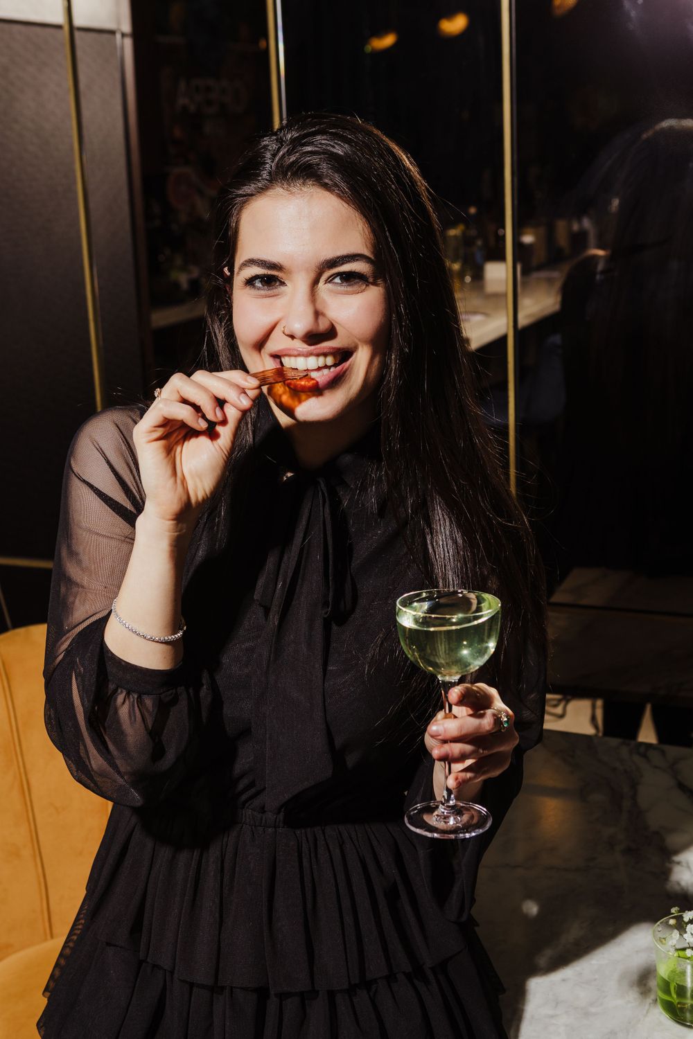 Person in black dress enjoying cocktail and appetizer in dimly lit restaurant setting.