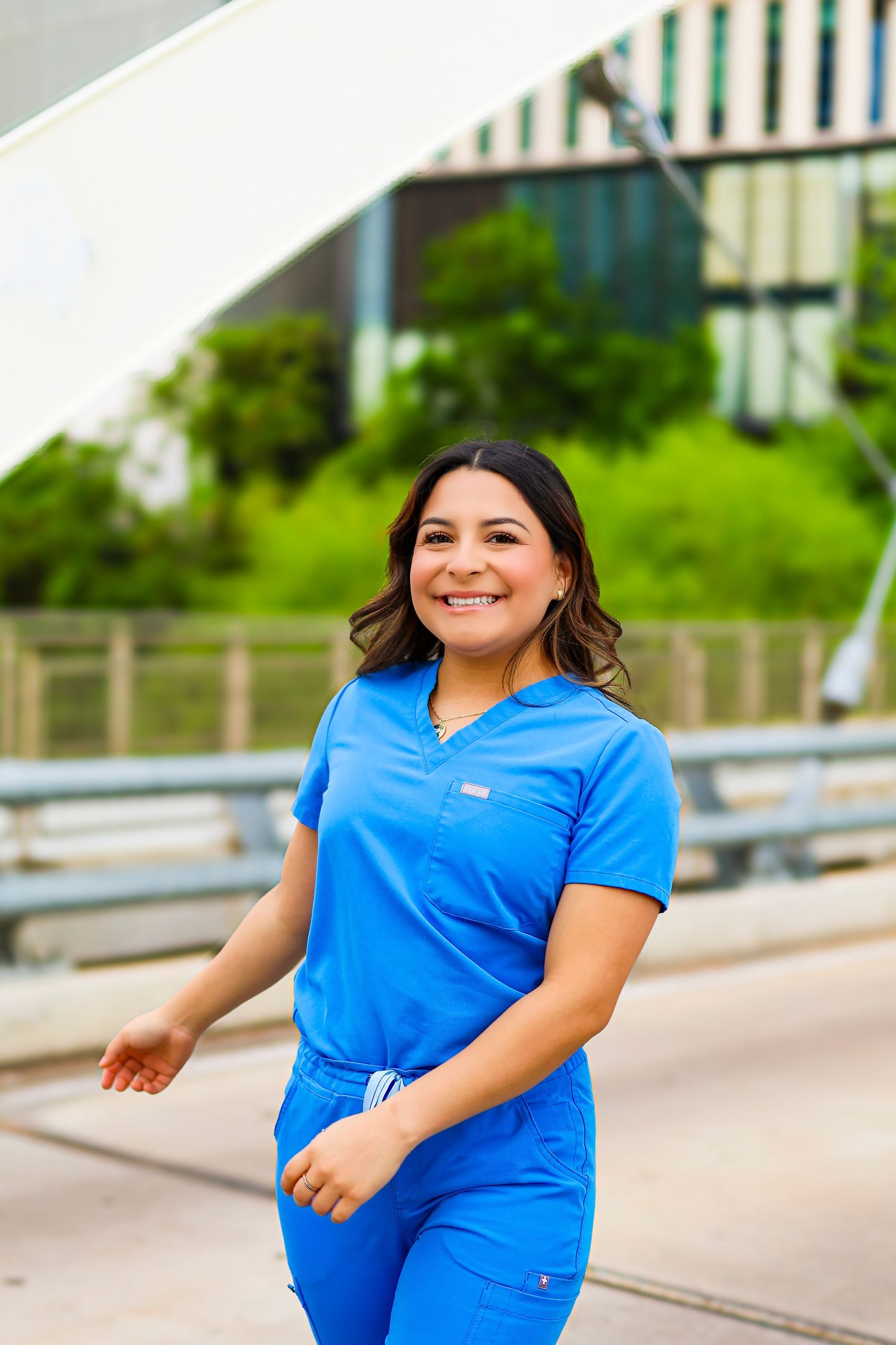 Medical professional in blue scrubs smiling outdoors with greenery in background.