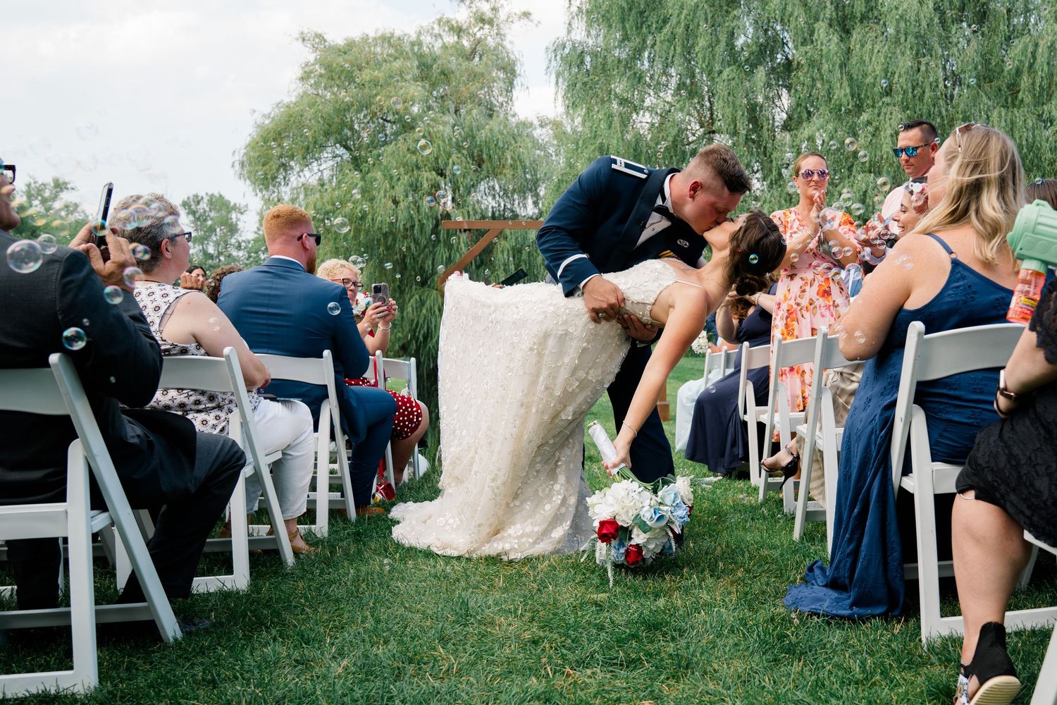 Wedding guests seated in white chairs on a lawn beneath willow trees during an outdoor ceremony.
