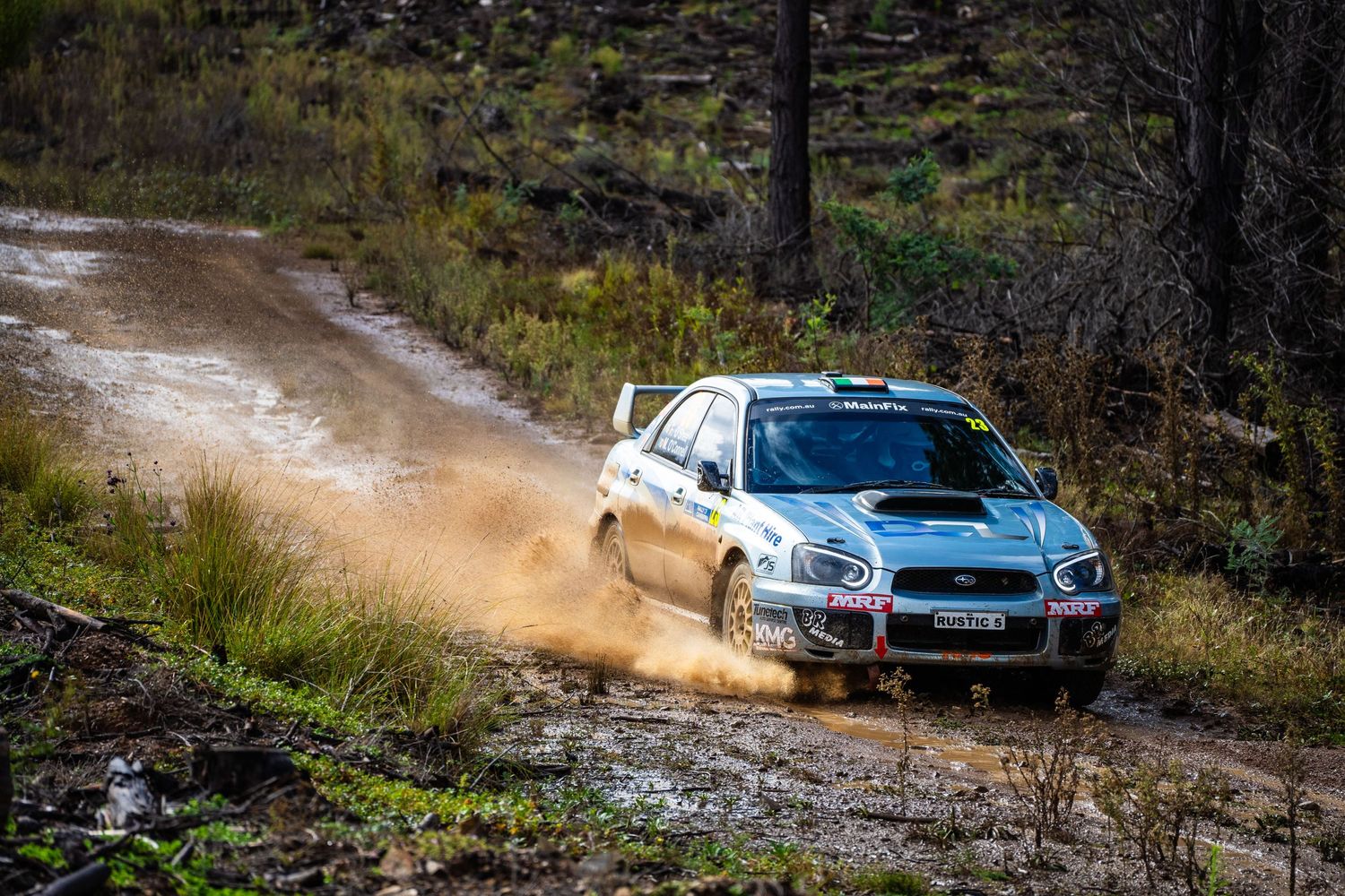 Rally car splashing through muddy terrain on forest rally course.