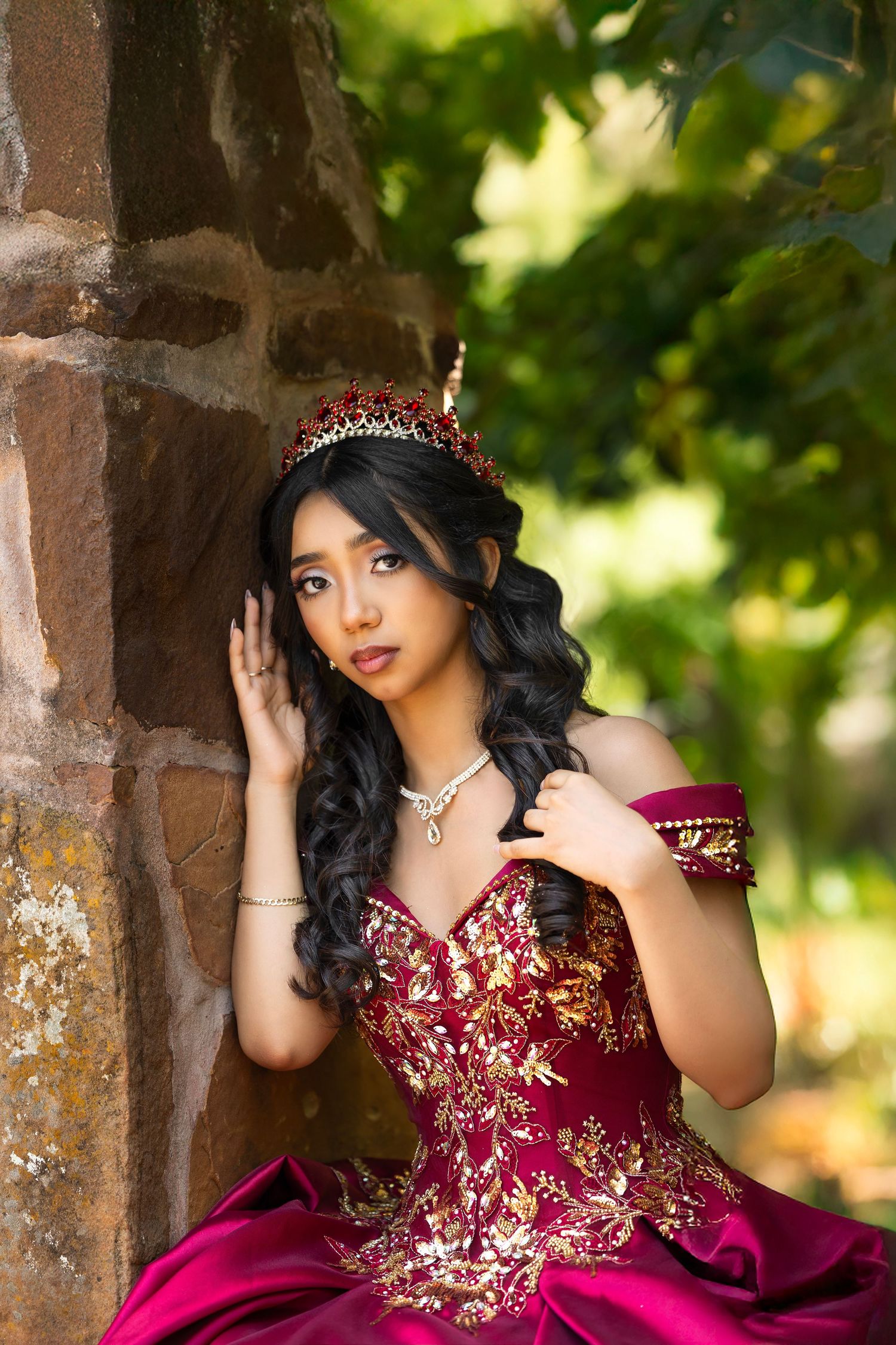 Elegant quinceanera portrait showcasing a burgundy and gold embellished dress with tiara in an outdoor garden setting.