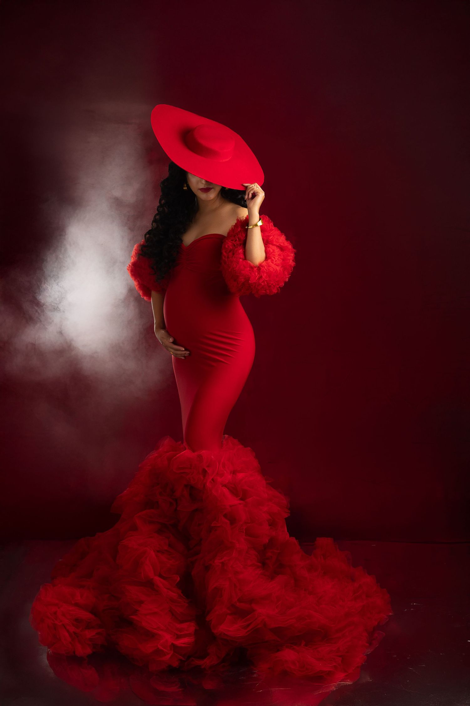 Dramatic fashion photo of a woman in a flowing red mermaid gown and matching hat posed against a dark smoky backdrop.