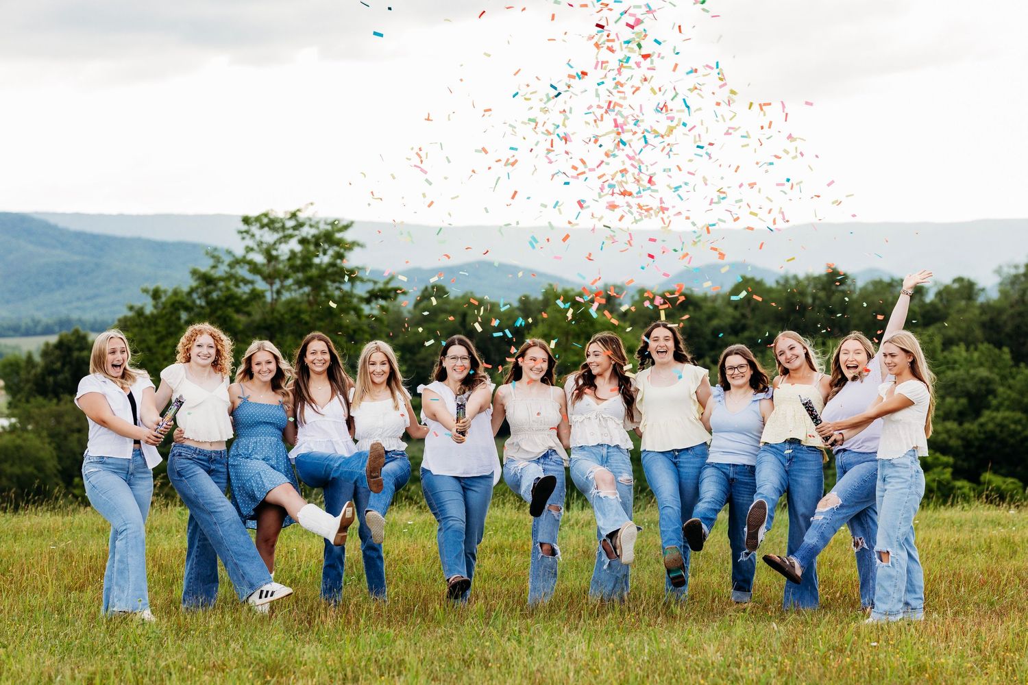 Group of women in white tops and jeans celebrating with colorful confetti in mountain meadow.