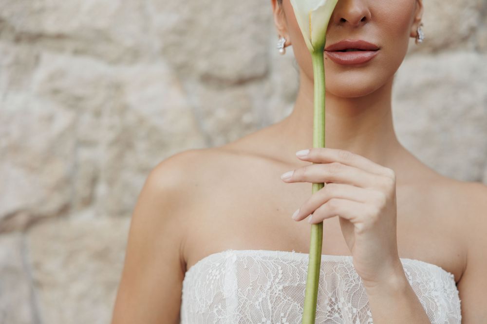 A bride in a white strapless wedding dress holds a single elegant calla lily flower against a stone wall background.