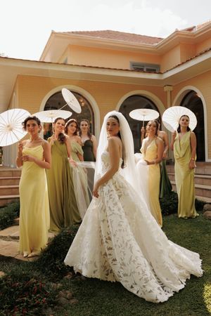A wedding party in yellow and sage green dresses poses with parasols in front of an elegant arched building.