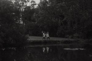 A black and white landscape photograph showing two figures reflected in a tranquil pond.