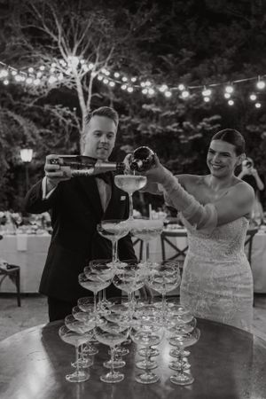 A champagne tower being filled at an outdoor evening celebration with string lights.