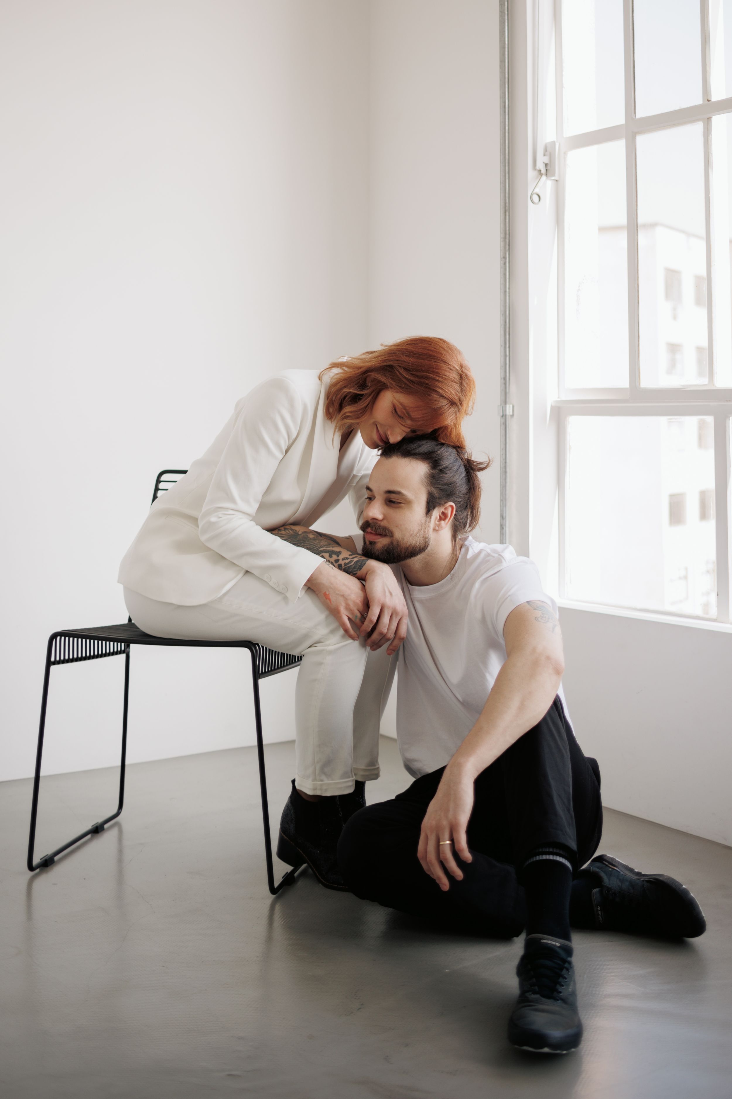 Two people in white clothing share an intimate moment near a bright window in a minimalist room setting.