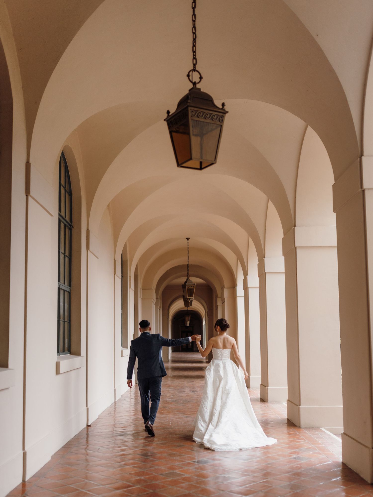 Bride and groom walking hand in hand through elegant arched corridor with hanging lanterns.