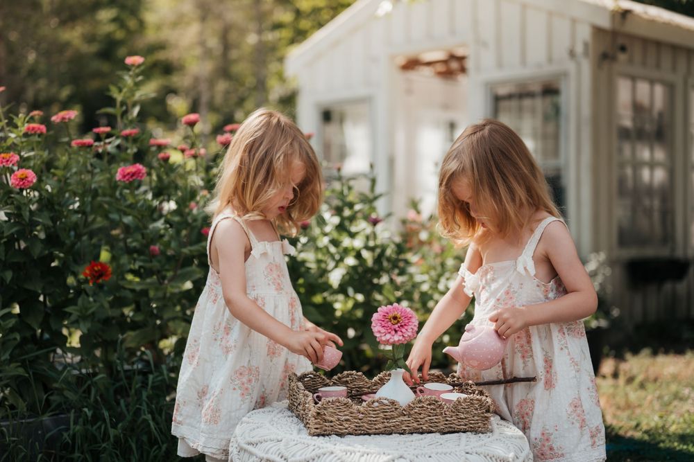 Two children play near a garden of virannt beautiful flowers with a white greenhouse in the background.
