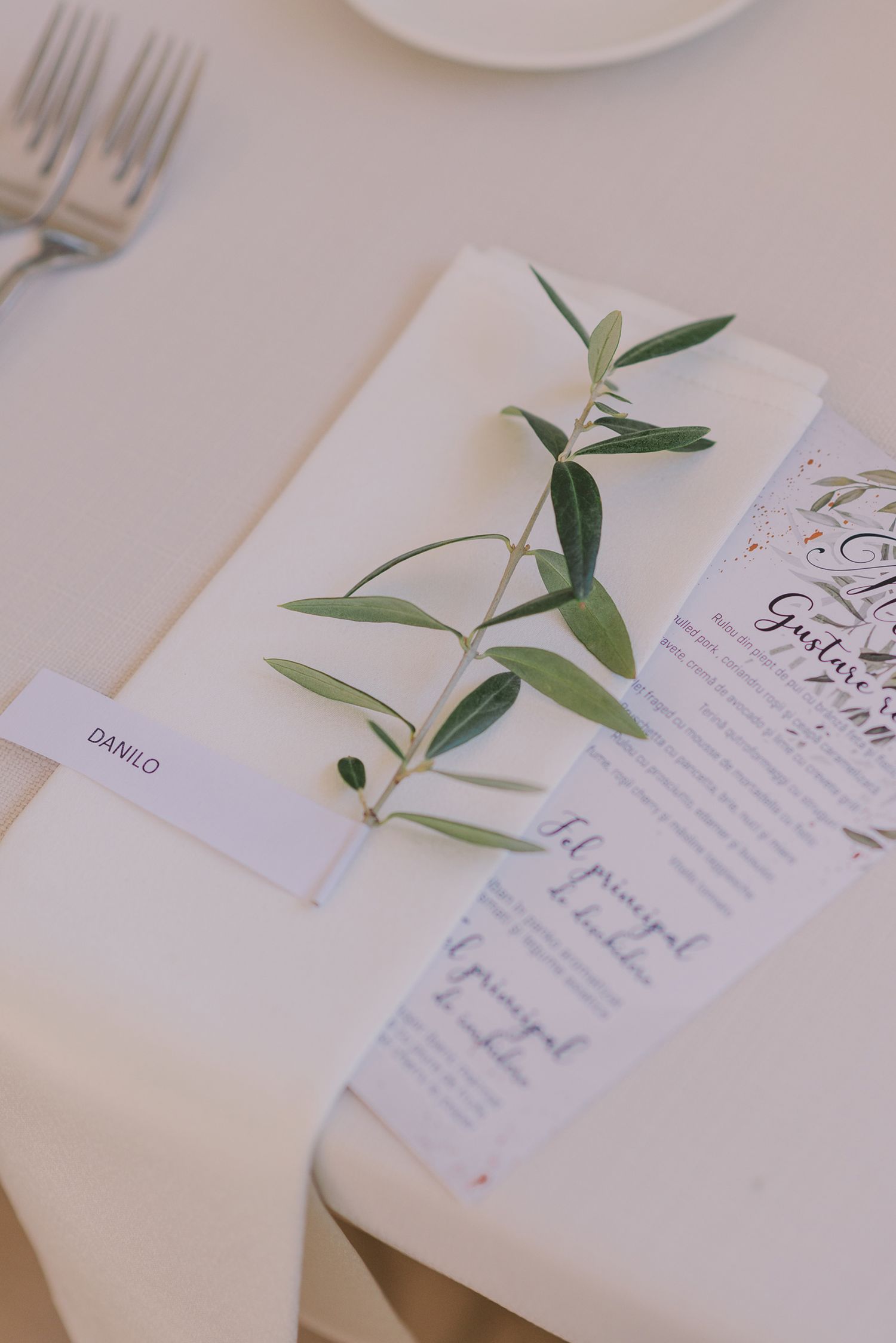 Olive branch place setting with menu card on white linen tablecloth.