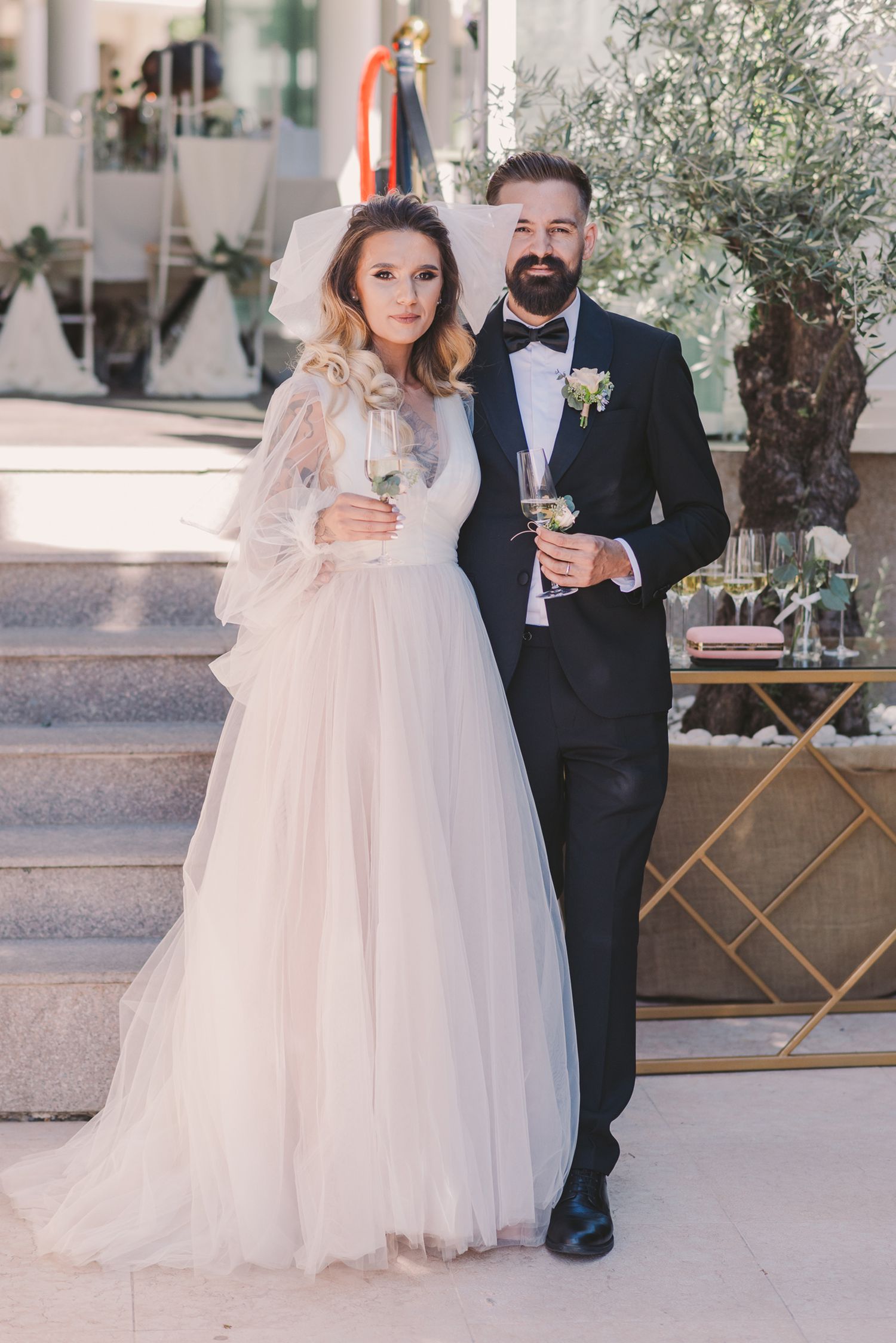 Wedding couple poses on steps with olive tree and champagne glasses.