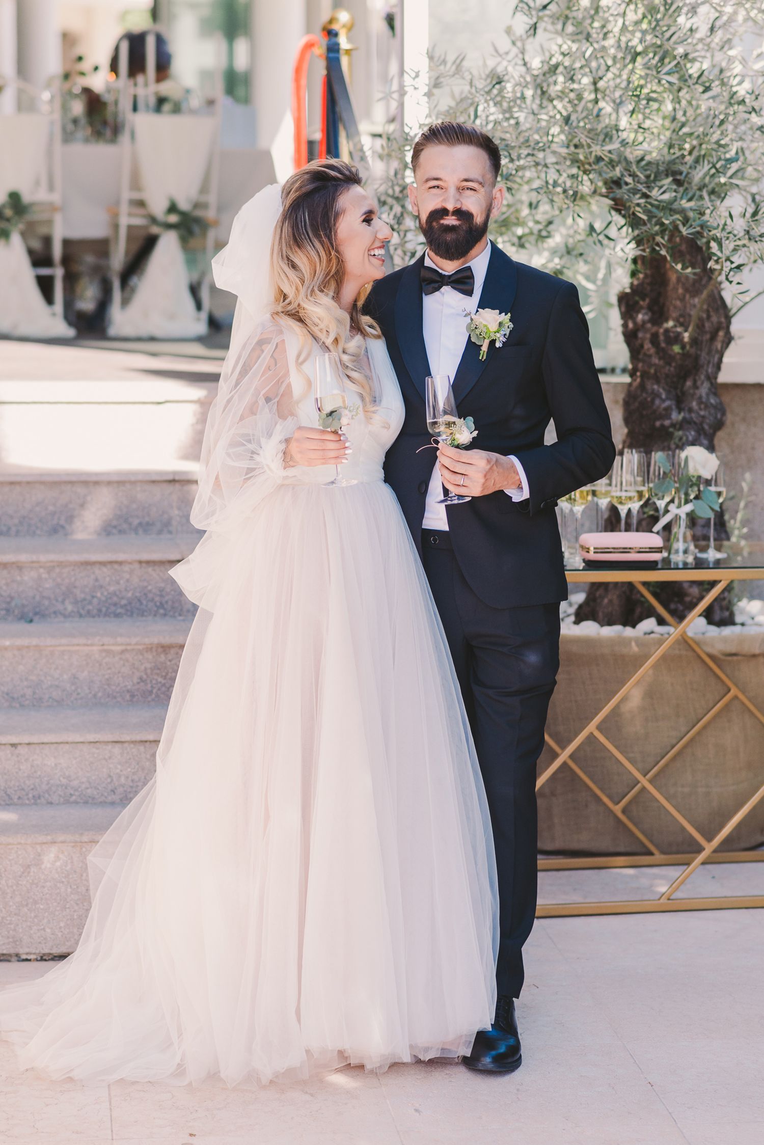 Newlyweds share intimate moment on outdoor steps with olive tree backdrop.