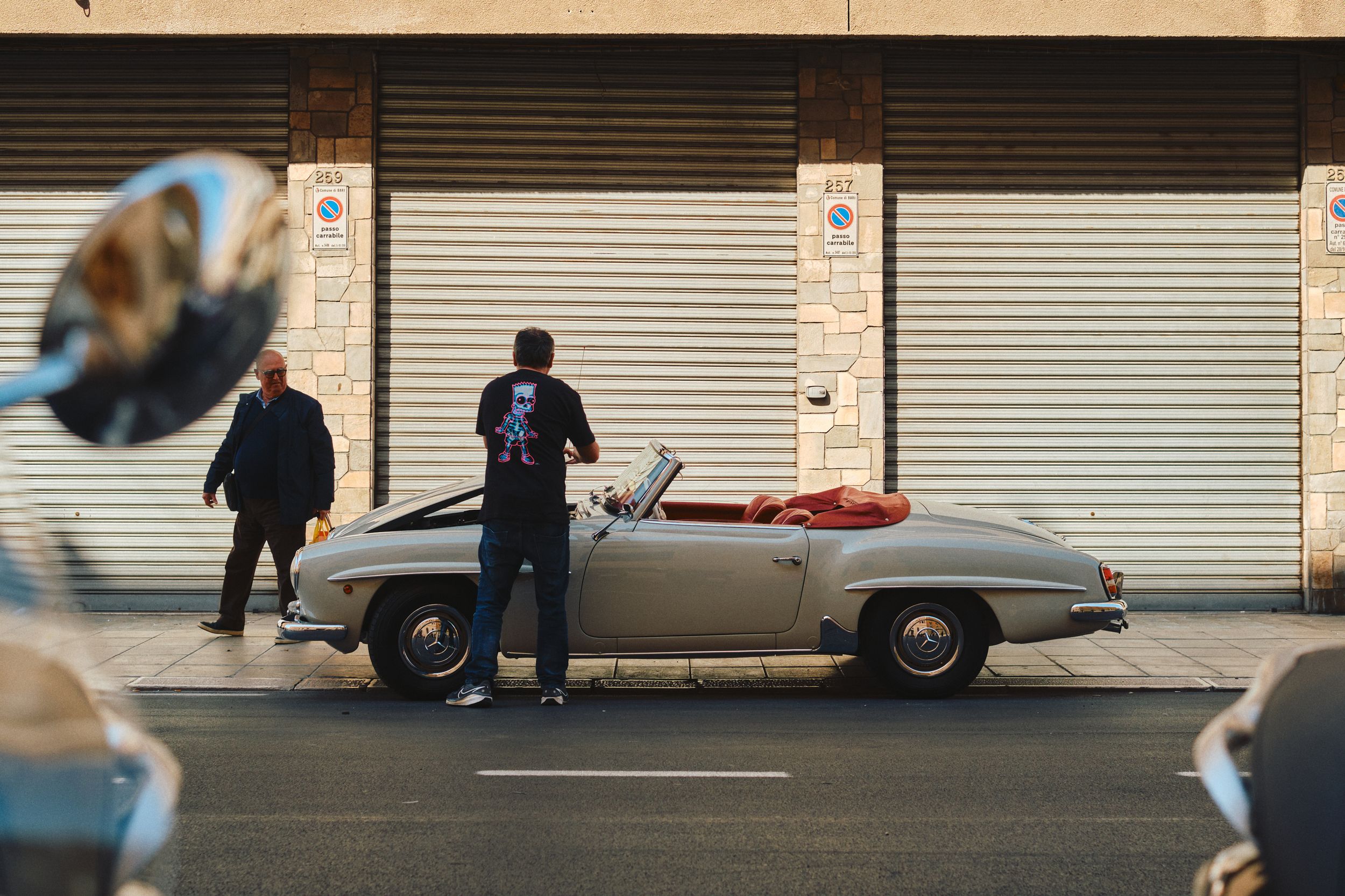 Classic convertible car parked outside closed storefront shutters on urban street.