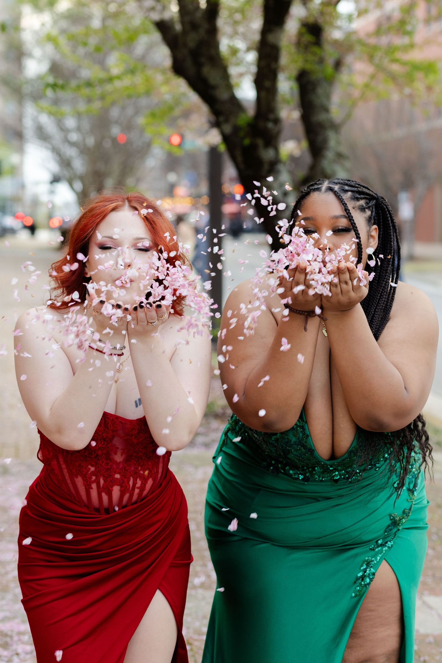 Two high school seniors in prom dresses blowing cherry blossom petals at camera.