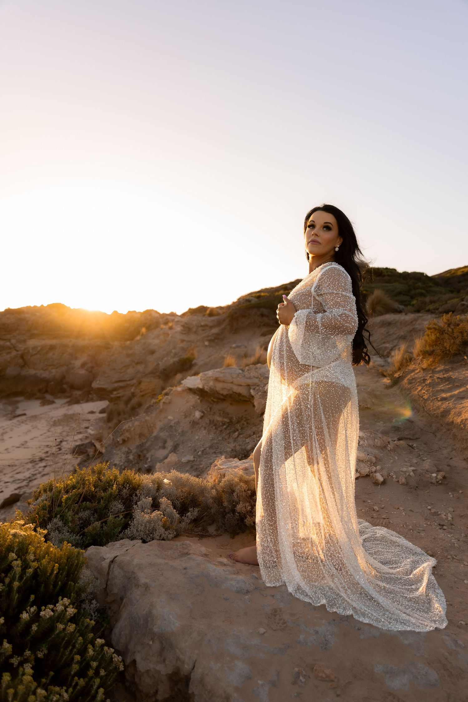 Pregnant woman in lace gown poses on rocky cliff at sunset.