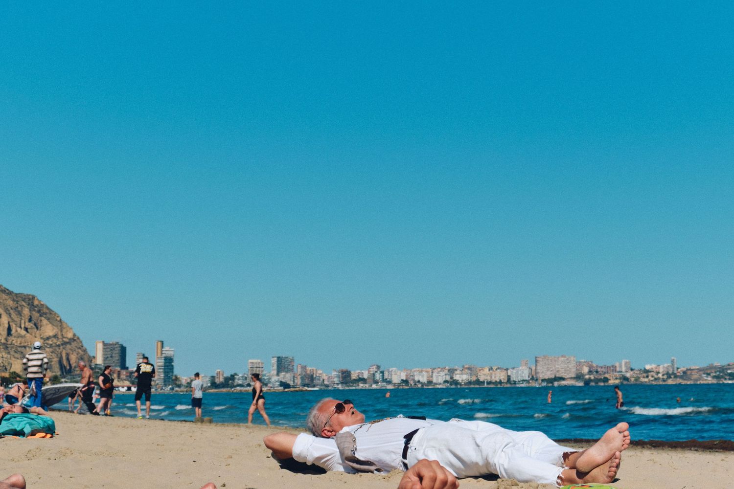People relaxing on sandy beach with coastal cityscape in background.