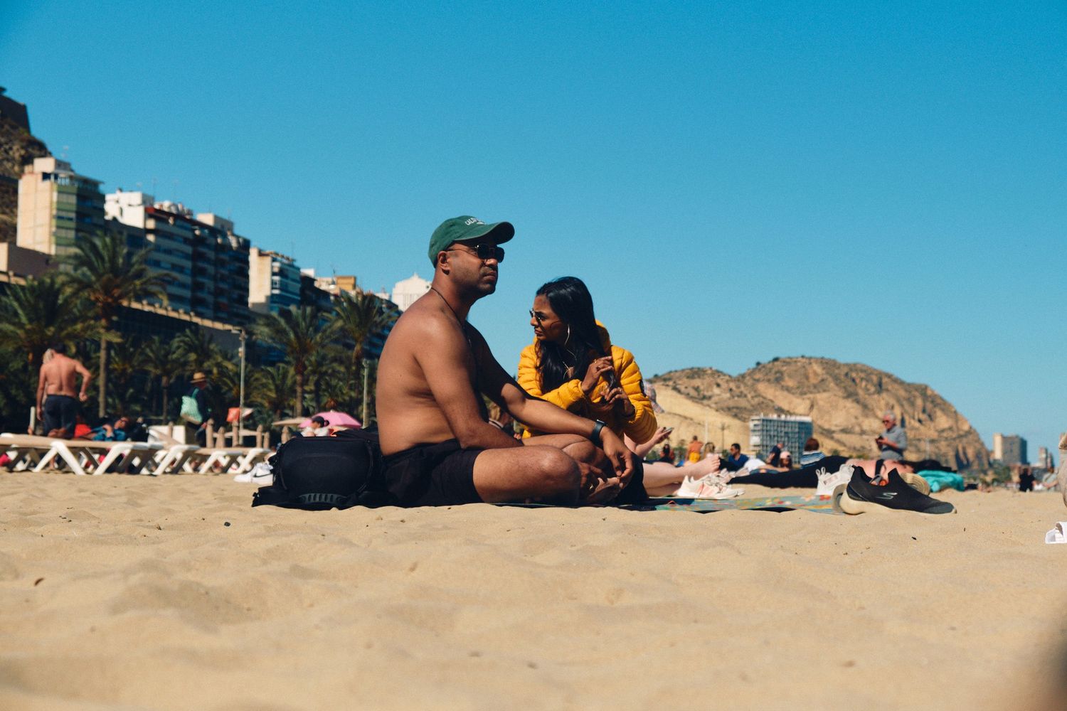 Beachgoers relax on a sandy shoreline with coastal buildings and mountains in the background.