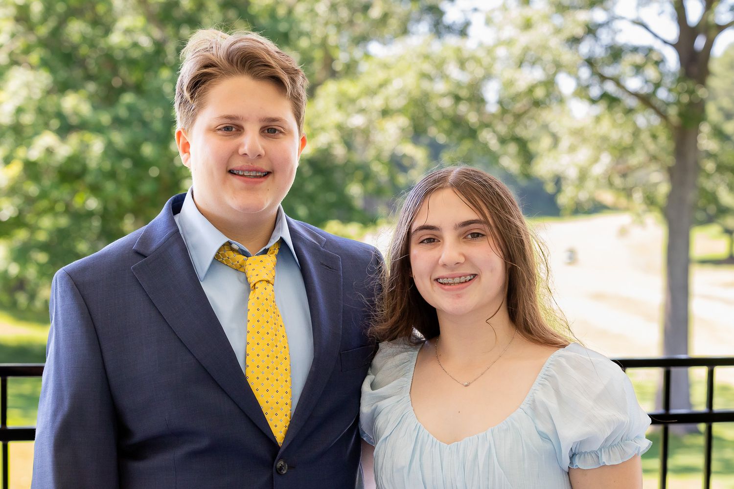 A young couple in formal wear poses outside on a sunny day against a blurred green background.