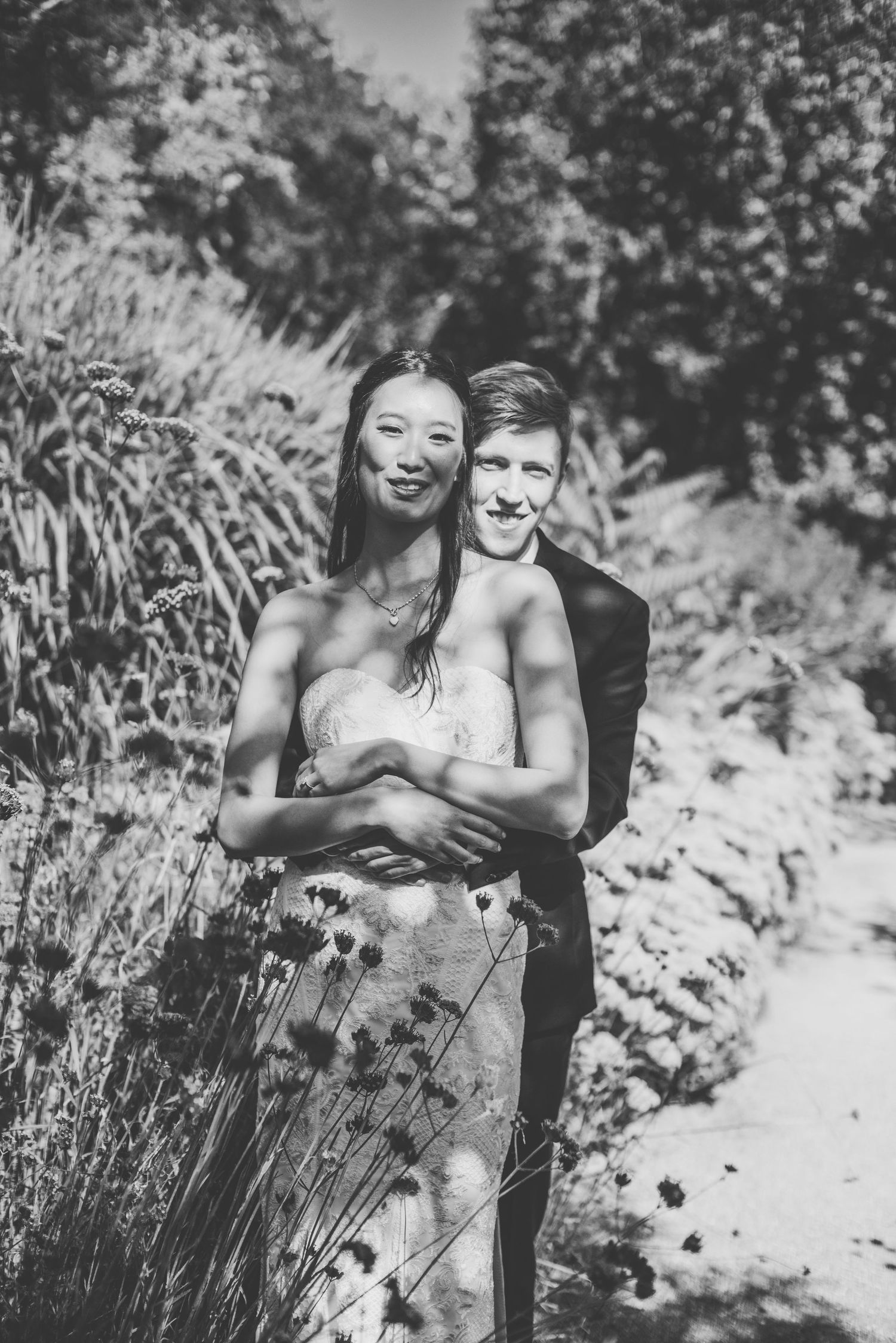 Black and white portrait of couple embracing among tall grasses in garden setting.