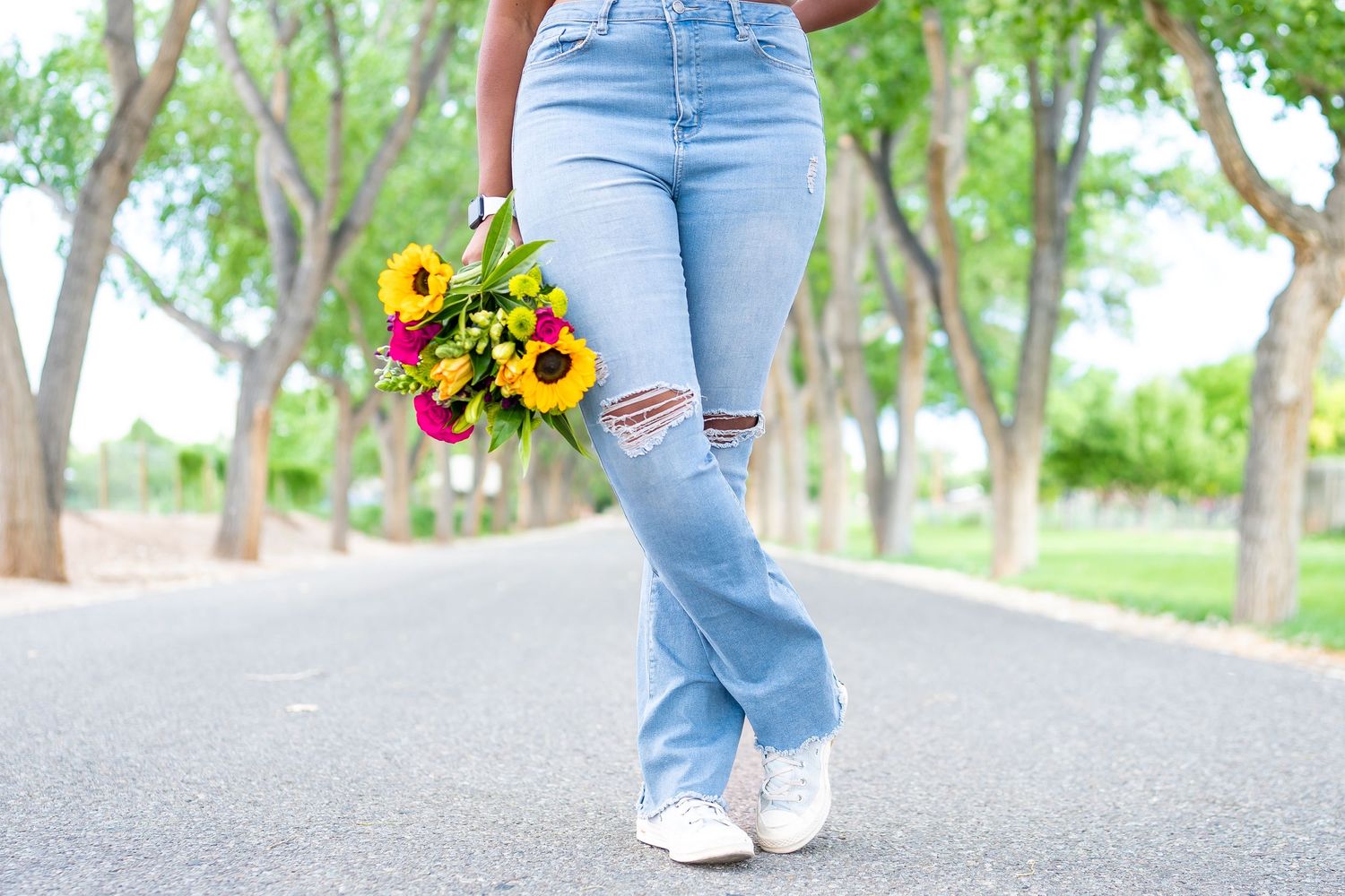 Ripped blue jeans and white sneakers with colorful sunflower bouquet on tree-lined path.