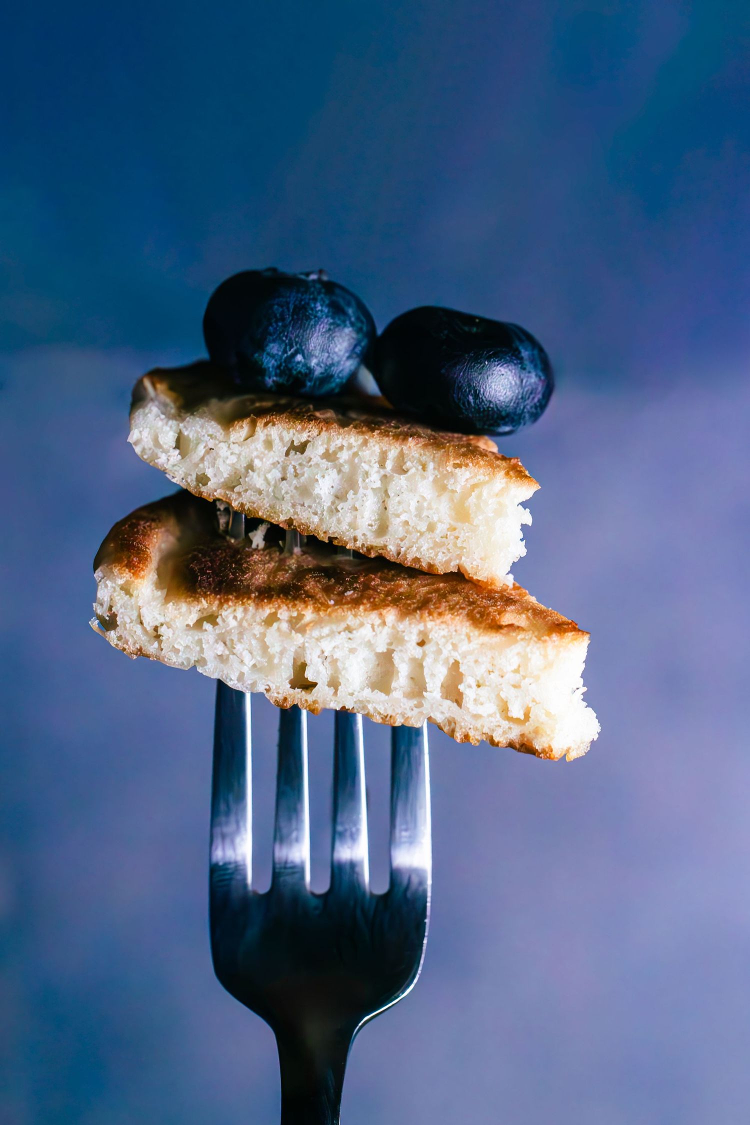 Two pieces of pancake with two blueberries stack on top of a stainless steel fork against a blue background.