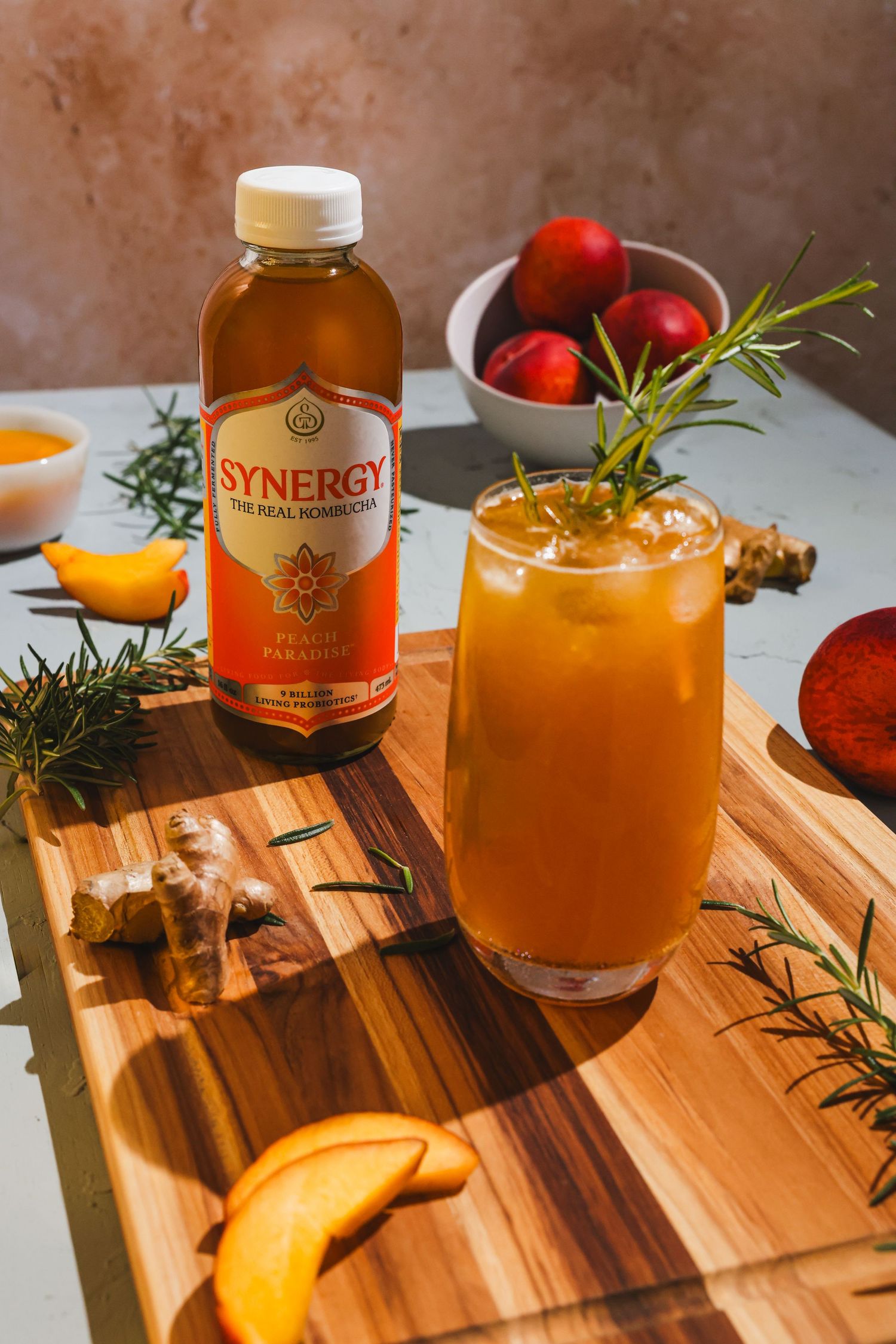 A bottle of Synergy kombucha next to a glass of peach mocktail garnished with fresh rosemary staged on a wooden cutting board.