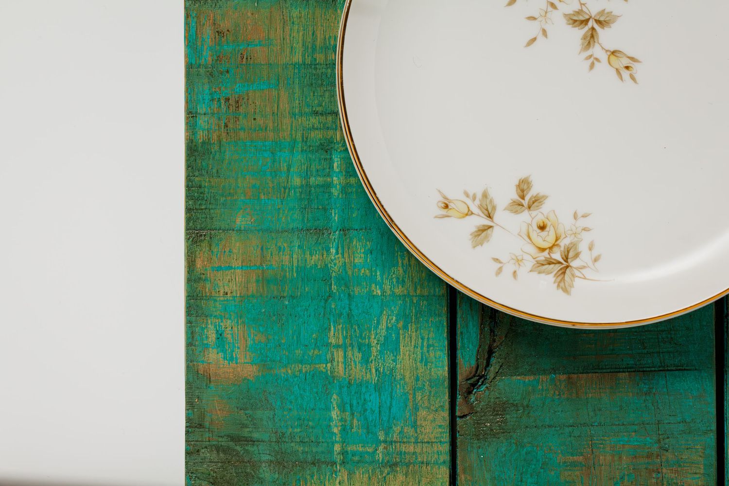 Vintage white porcelain plate with delicate gold floral details rests on a distressed turquoise wooden surface on a white table.