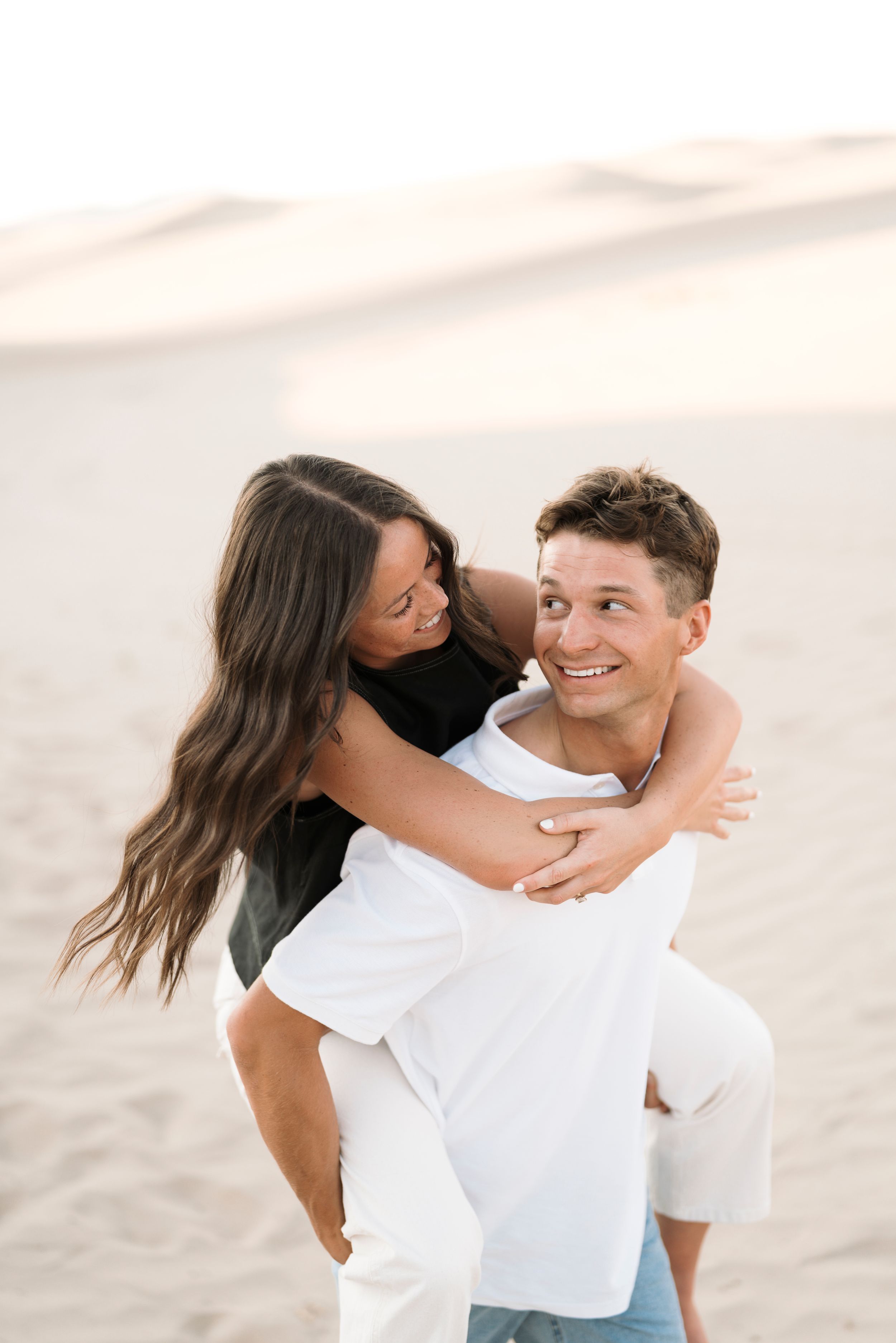 A happy couple shares a piggyback ride on sandy dunes, smiling and embracing in casual white and black outfits.