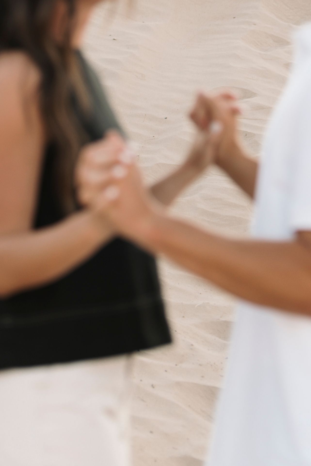 Two people holding hands on a sandy beach, one wearing black and one in white.