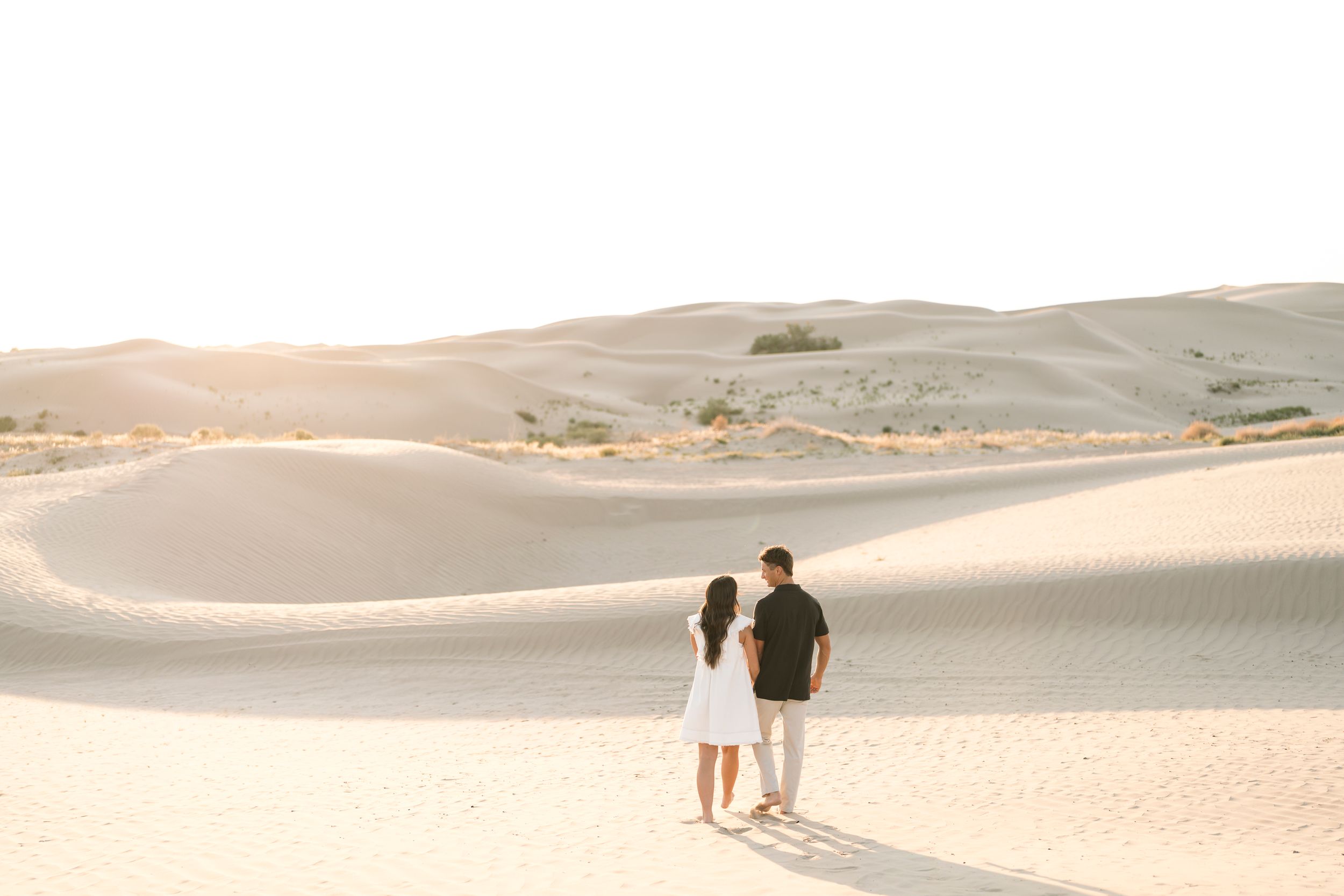 Couple holding hands walking through vast sandy desert dunes at golden hour sunset.