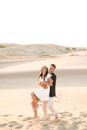 Couple laughing and embracing barefoot on sandy desert dunes, woman in white dress, man in black shirt and white pants.