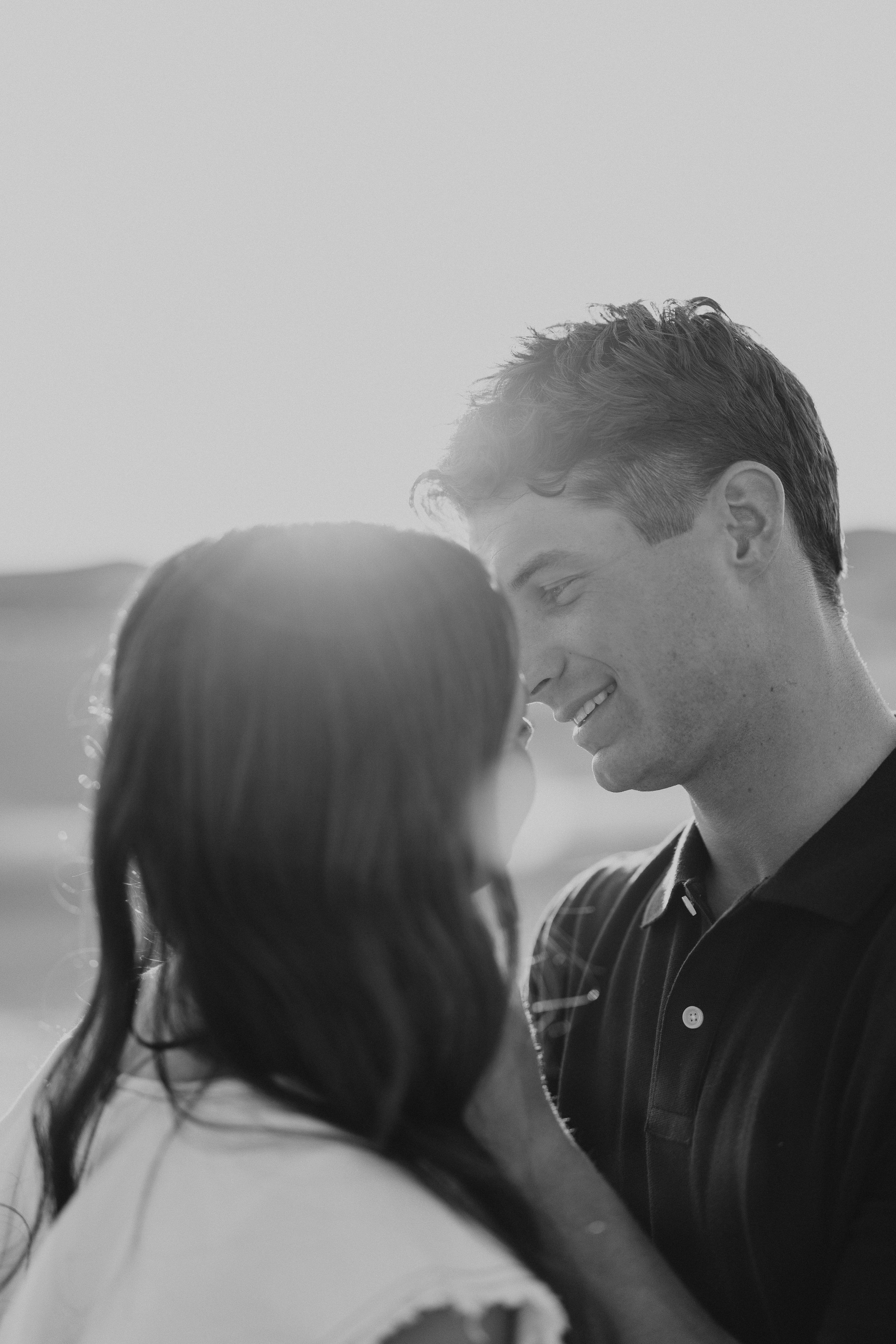Black and white photo of a smiling couple sharing a romantic moment, backlit by soft sunlight outdoors.