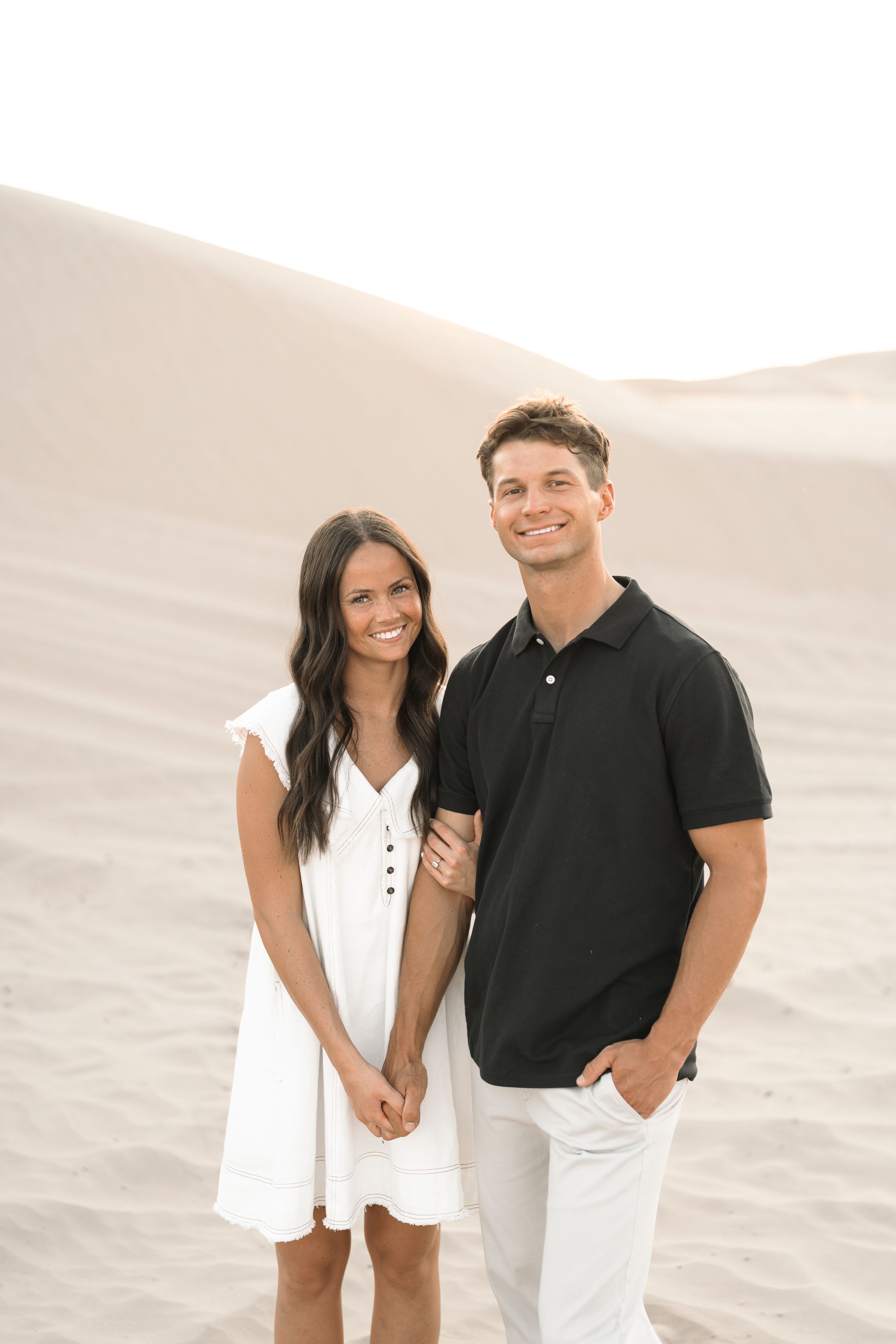 A smiling couple poses on sandy dunes, woman in white dress, man in black polo and white pants.