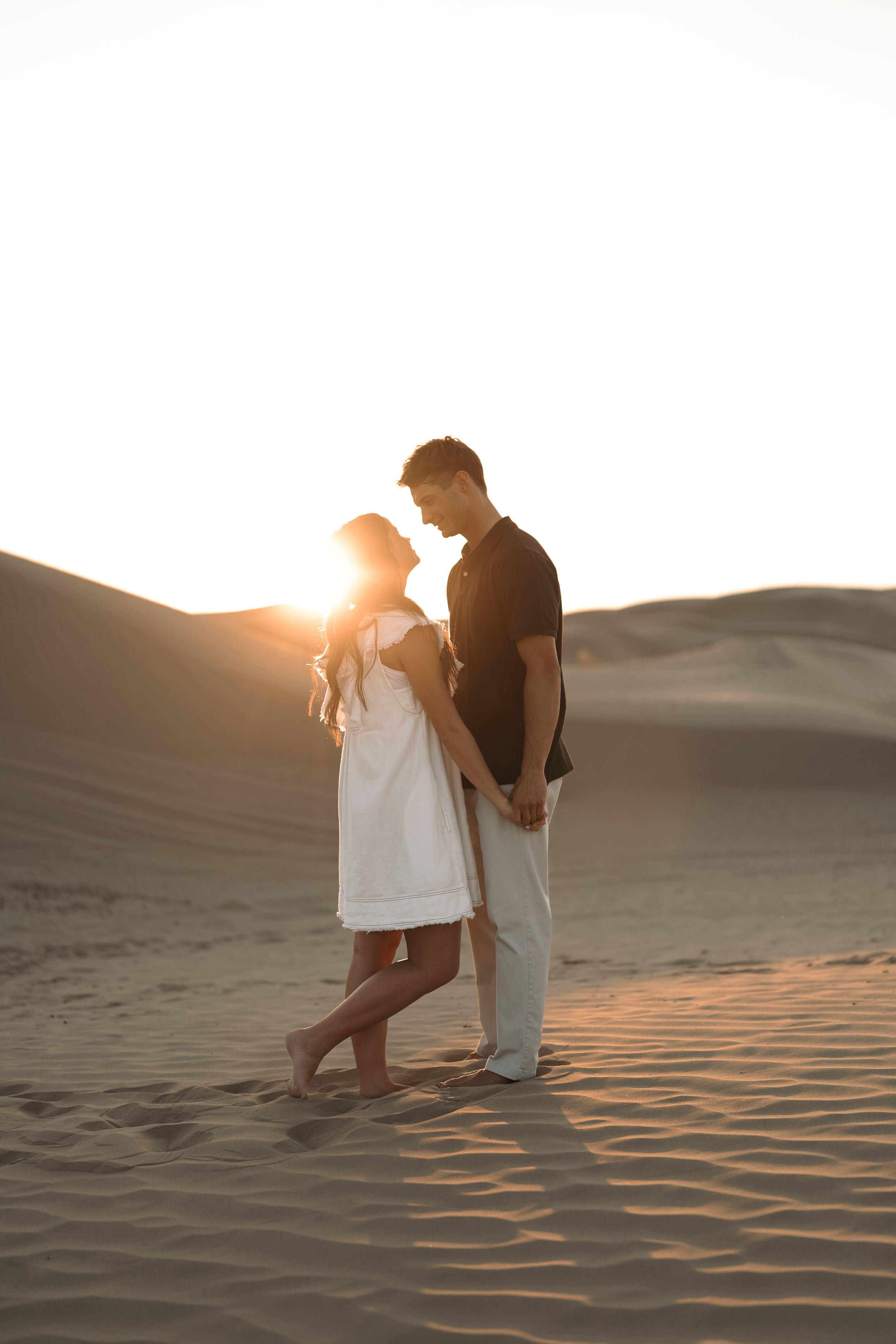 Romantic couple holding hands at sunset on desert sand dunes, silhouetted against golden backlight.